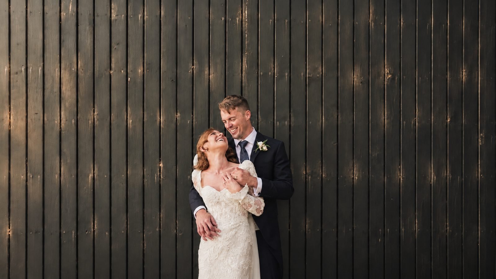 Couple laughing together against a dark timber barn wall